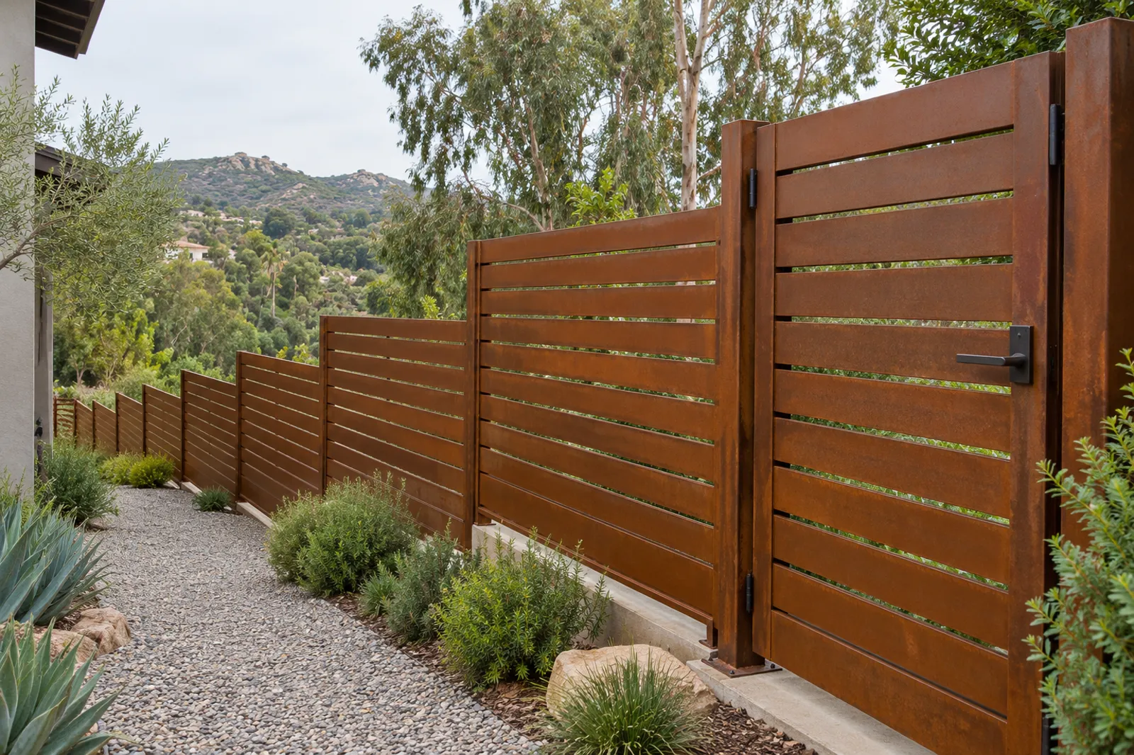 Horizontal Cor-Ten steel slat fence and matching walk gate stepping along a gravel path, hills and eucalyptus behind
