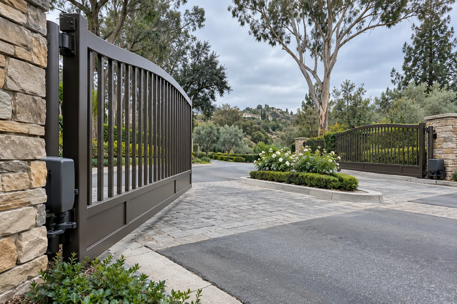 Black steel swing gate between stone pillars at a gated community entry, flower-planted median and eucalyptus trees behind