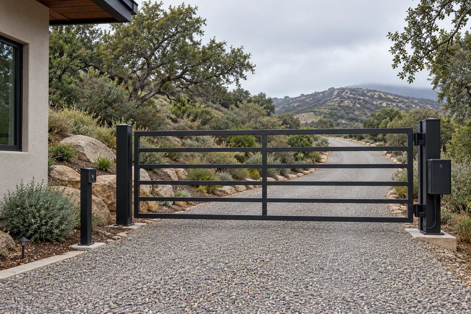 Black steel ranch-style tube gate across a long gravel drive, operator box on the post and a key switch post, chaparral hills