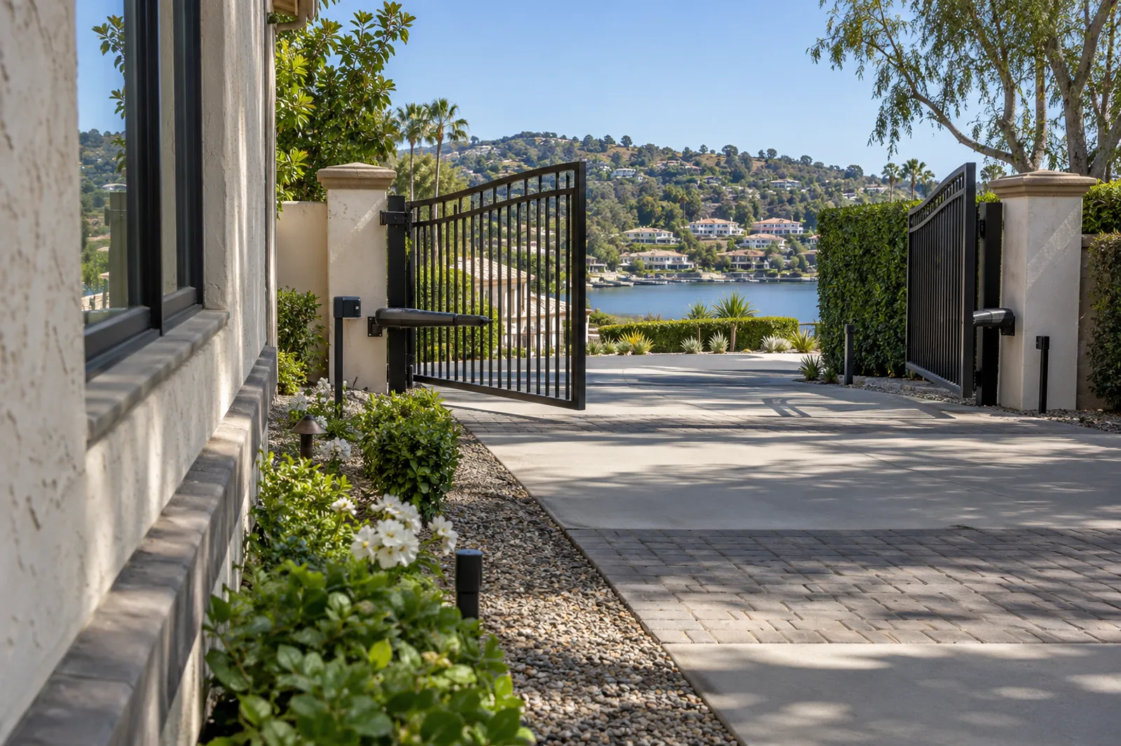 Automated black steel swing gates standing open at a community drive above a lake, photo eye posts on both sides