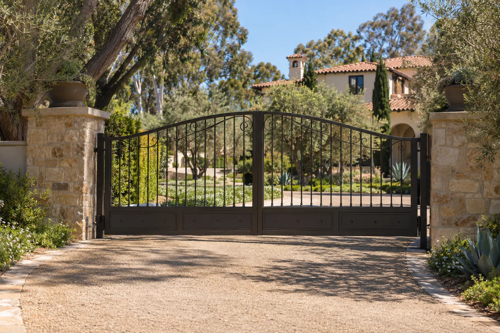 Arched black steel dual swing driveway gate between cut-stone pillars, crushed-granite drive leading to a Spanish-style estate